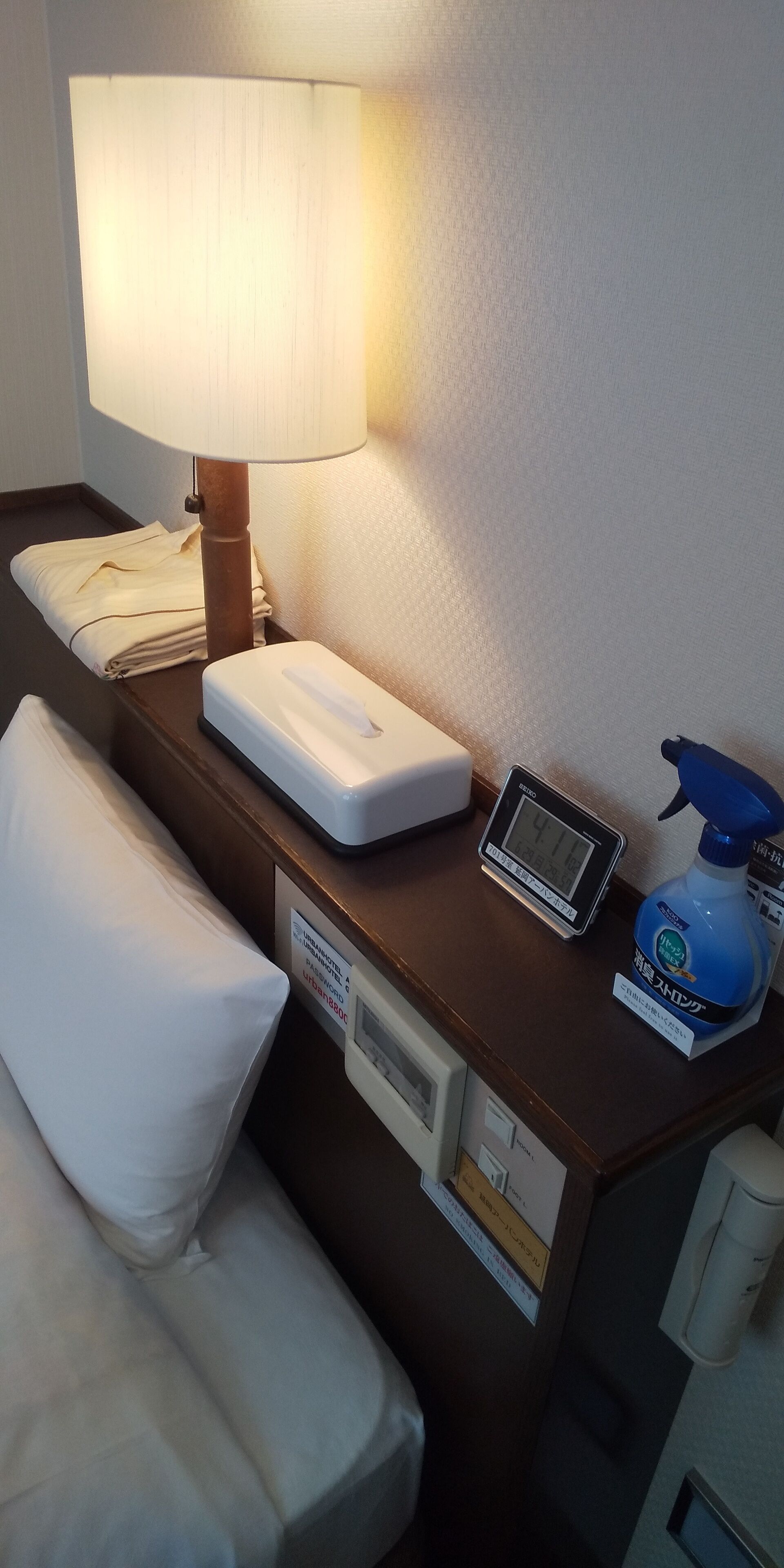 Habitació d'hotel amb una taula de nit, una làmpada, un rellotge digital, un borrifador i un coixí.