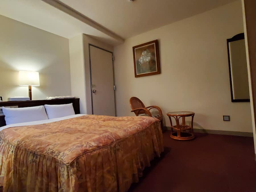 Quarto de hotel com cama, mesa de cabeceira com luminária, uma cadeira, uma mesinha e um quadro na parede.