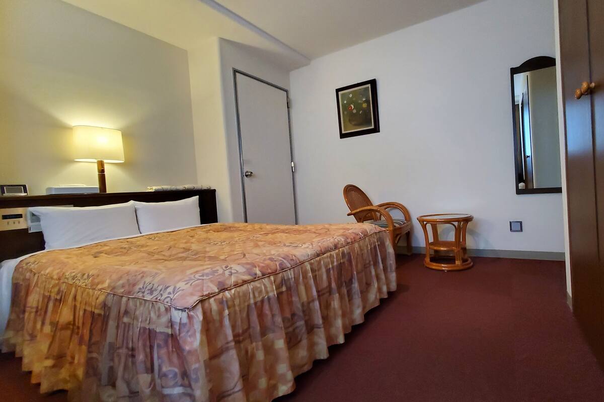 Une chambre d’hôtel avec un lit, une chaise, une petite table, une lampe, un tableau et un miroir.