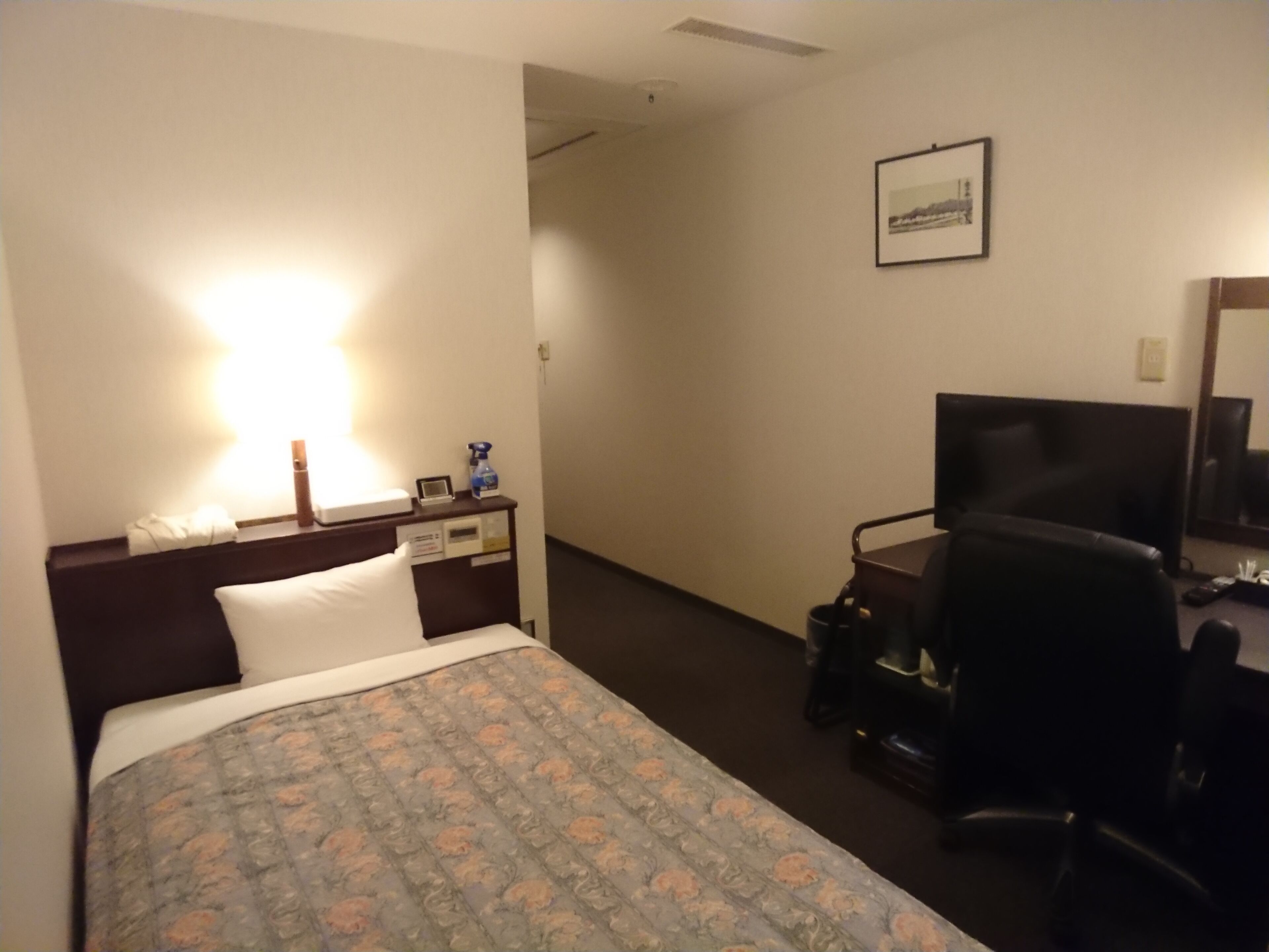 Una habitación de hotel con cama, escritorio, silla, lámpara de escritorio, mesita de noche y un cuadro en la pared.