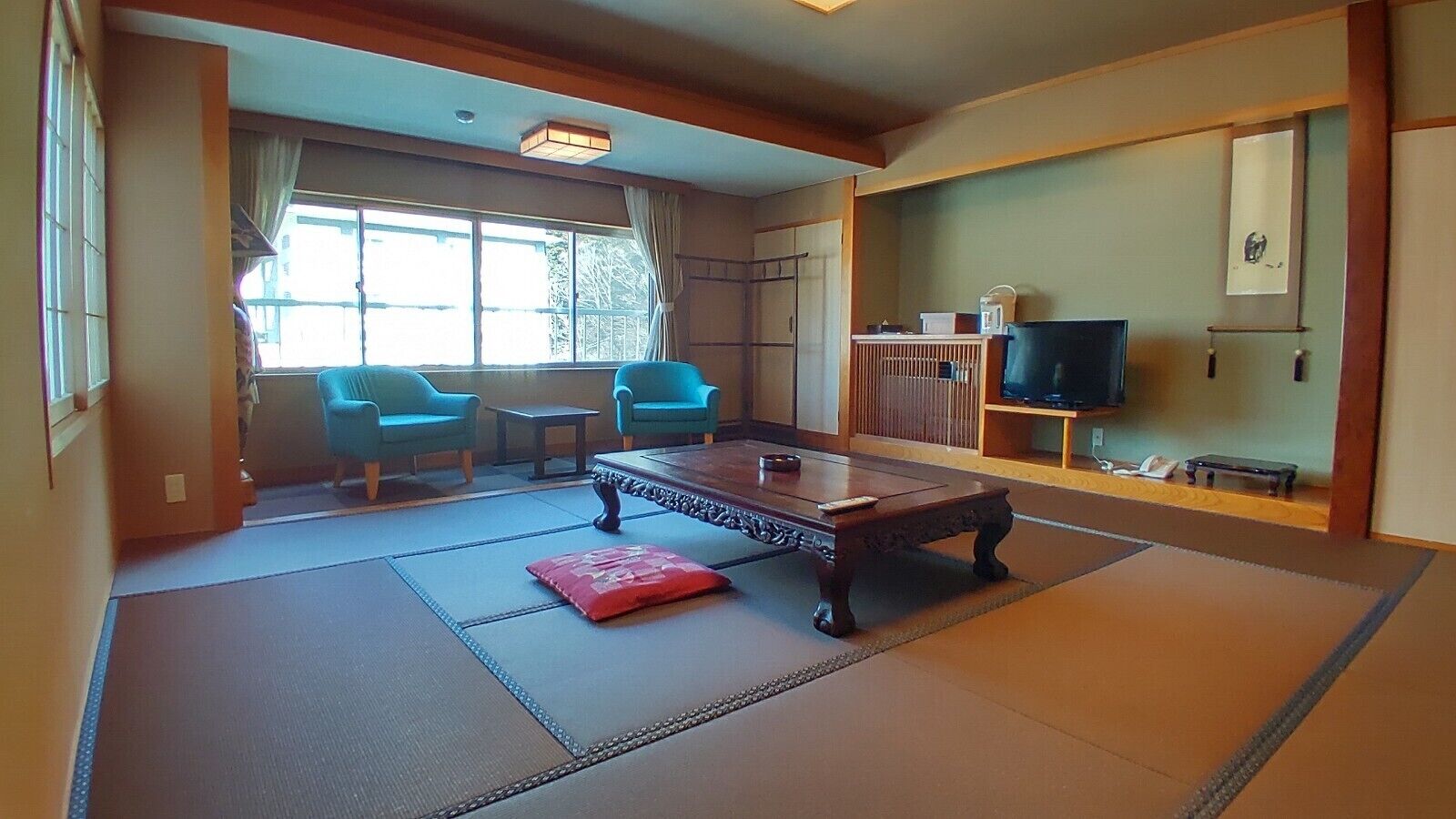 Una habitación tradicional japonesa con suelo de tatami, una mesa de madera baja, dos sillas azules, un televisor y una puerta corredera.