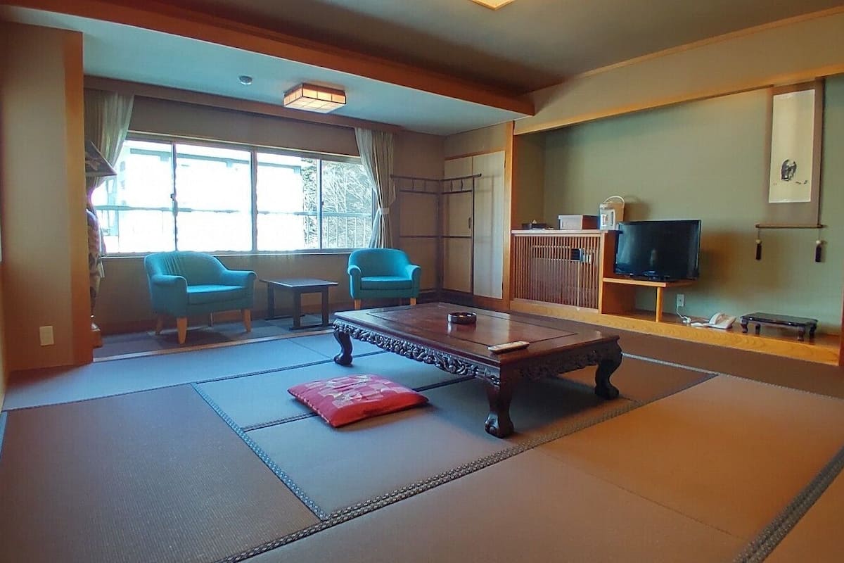 Ein traditioneller japanischer Raum mit Tatami-Boden, einem niedrigen Holztisch, zwei blauen Stühlen, einem Fernseher und einer Schiebetür.