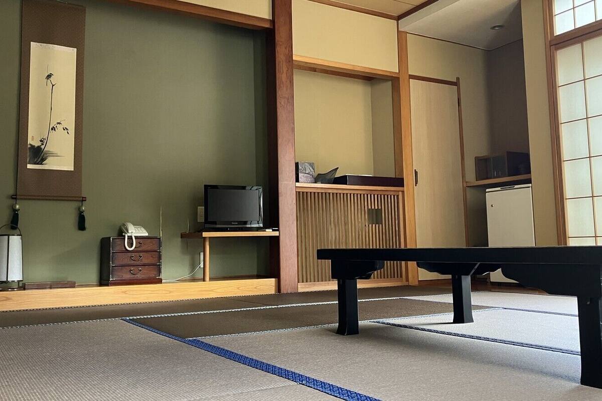 Ein Raum mit Tatami-Matten, einem niedrigen Tisch, einem Fernseher auf einem Holzständer und einer Schiebetür.