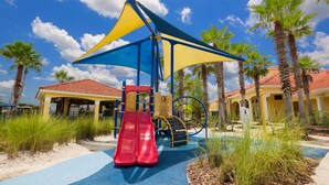 A playground with a red slide and a blue and yellow canopy, surrounded by palm trees and a building with a red-tiled roof.