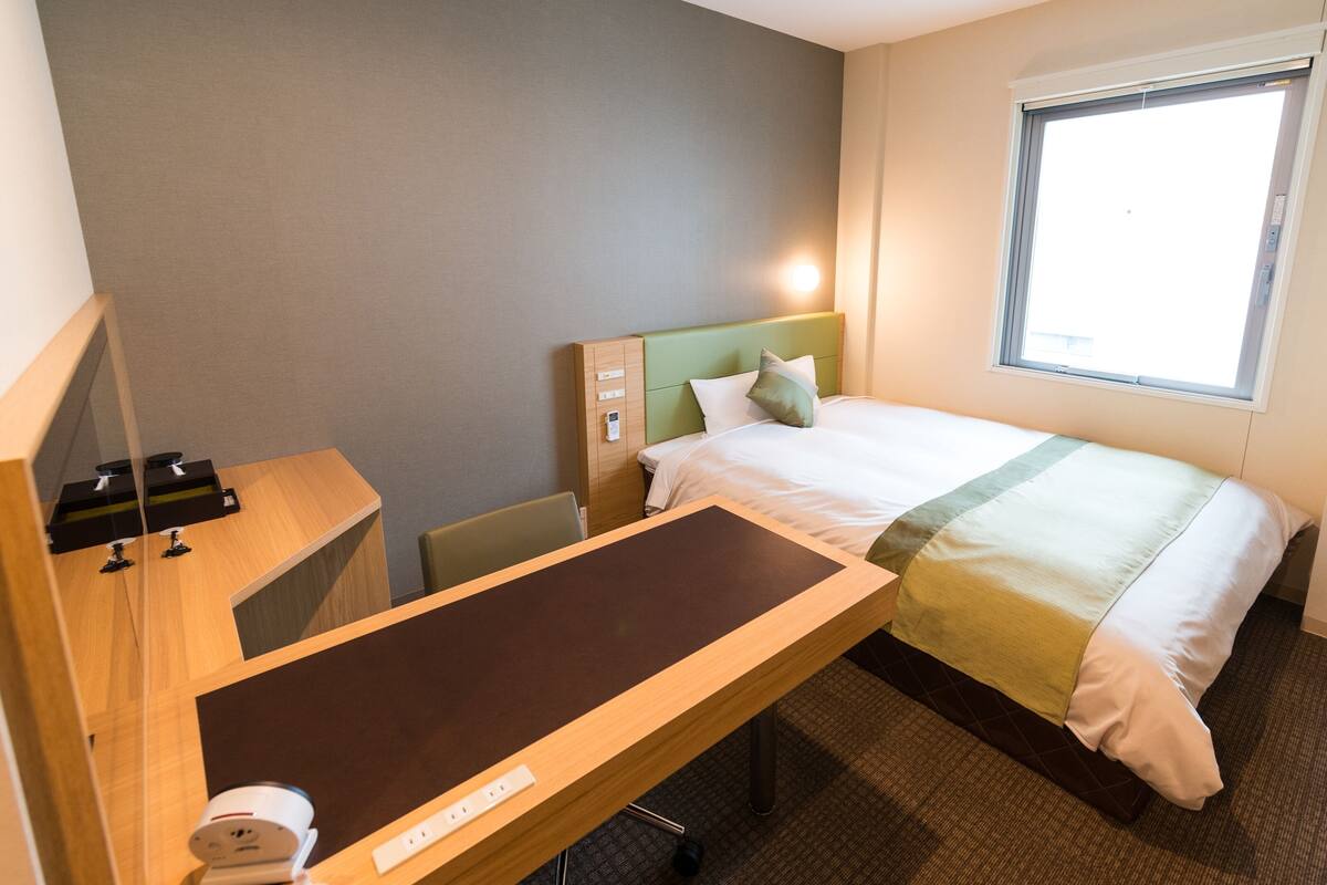 A hotel room with a bed, a desk with a telephone, a chair, and a window.