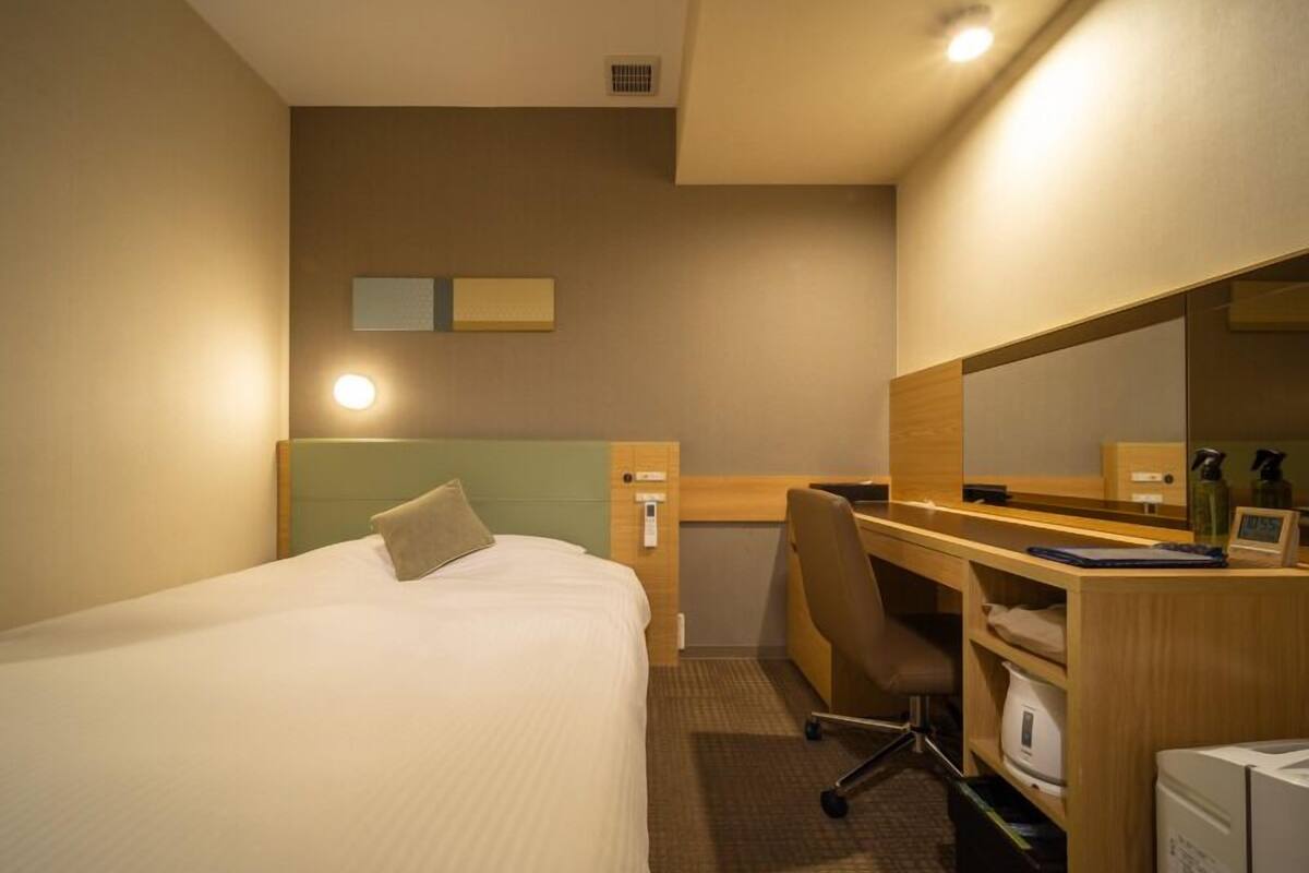 A hotel room with a bed, a desk, a chair, a mirror, a clock, and a refrigerator.