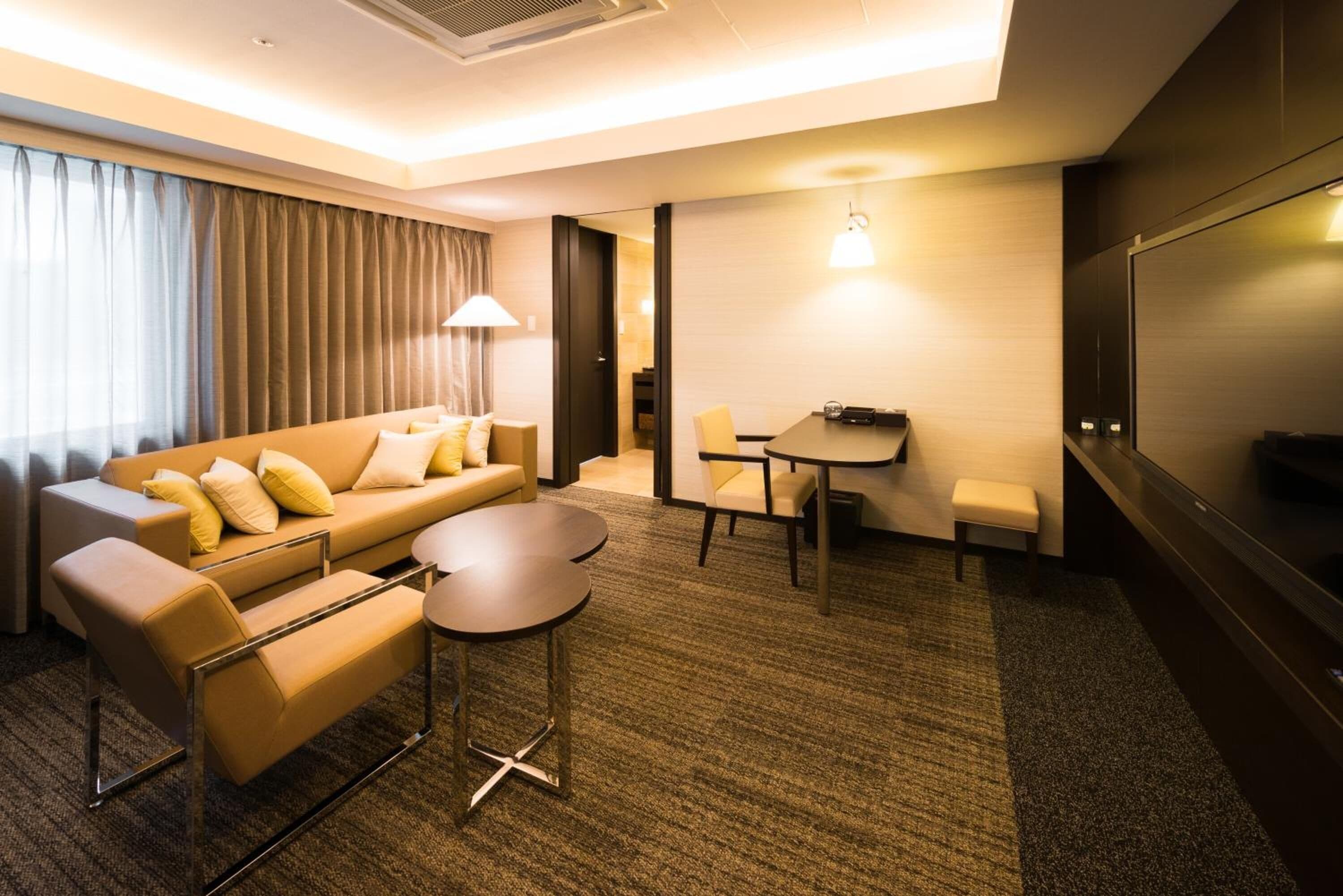 A modern hotel room with a sofa, two armchairs, a small round table, a desk, and a TV.