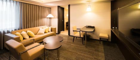 A modern hotel room with a sofa, two armchairs, a small round table, a desk, and a TV.