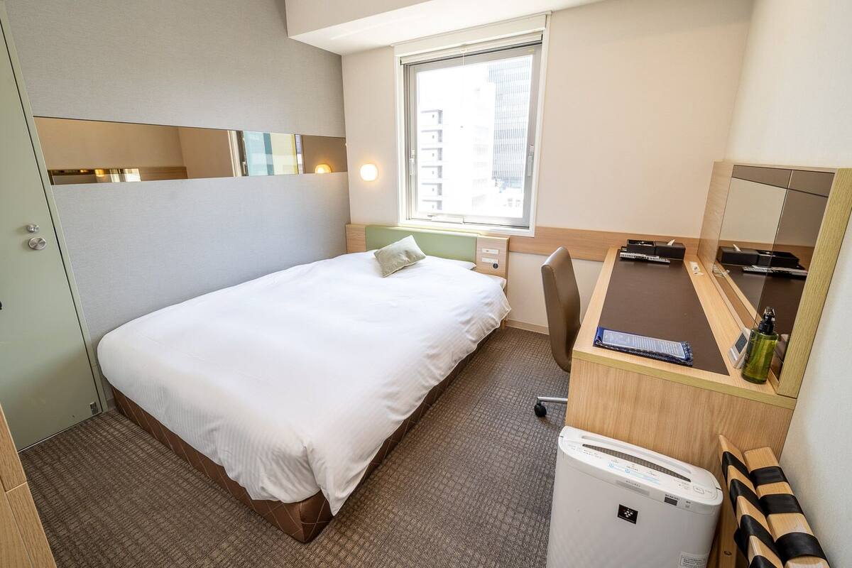 A hotel room with a bed, a desk, a mirror, a window, and a small air conditioning unit.