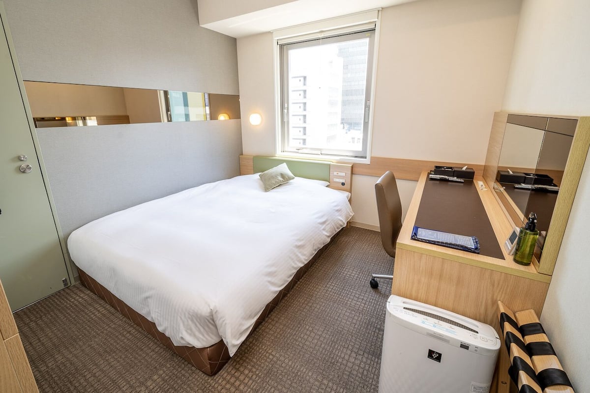 Habitación de hotel con cama, escritorio, espejo, ventana y unidad de aire acondicionado pequeña.