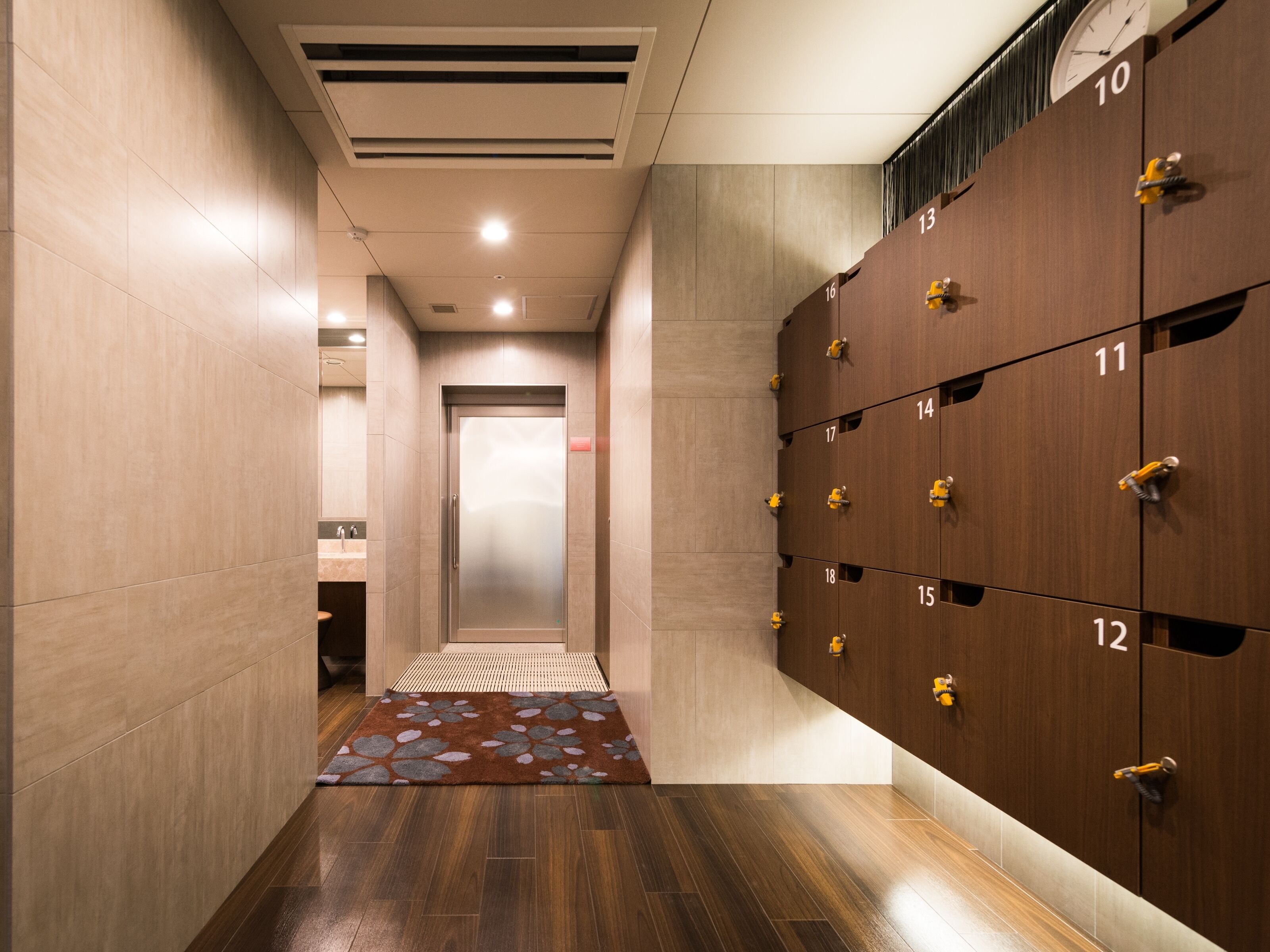 A modern hotel lobby with a central elevator, wooden lockers numbered 10 to 18, and a patterned rug on the floor.