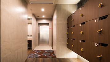 En moderne hotellobby med en central elevator, træskabe nummereret fra 10 til 18, og et mønstret tæppe på gulvet.