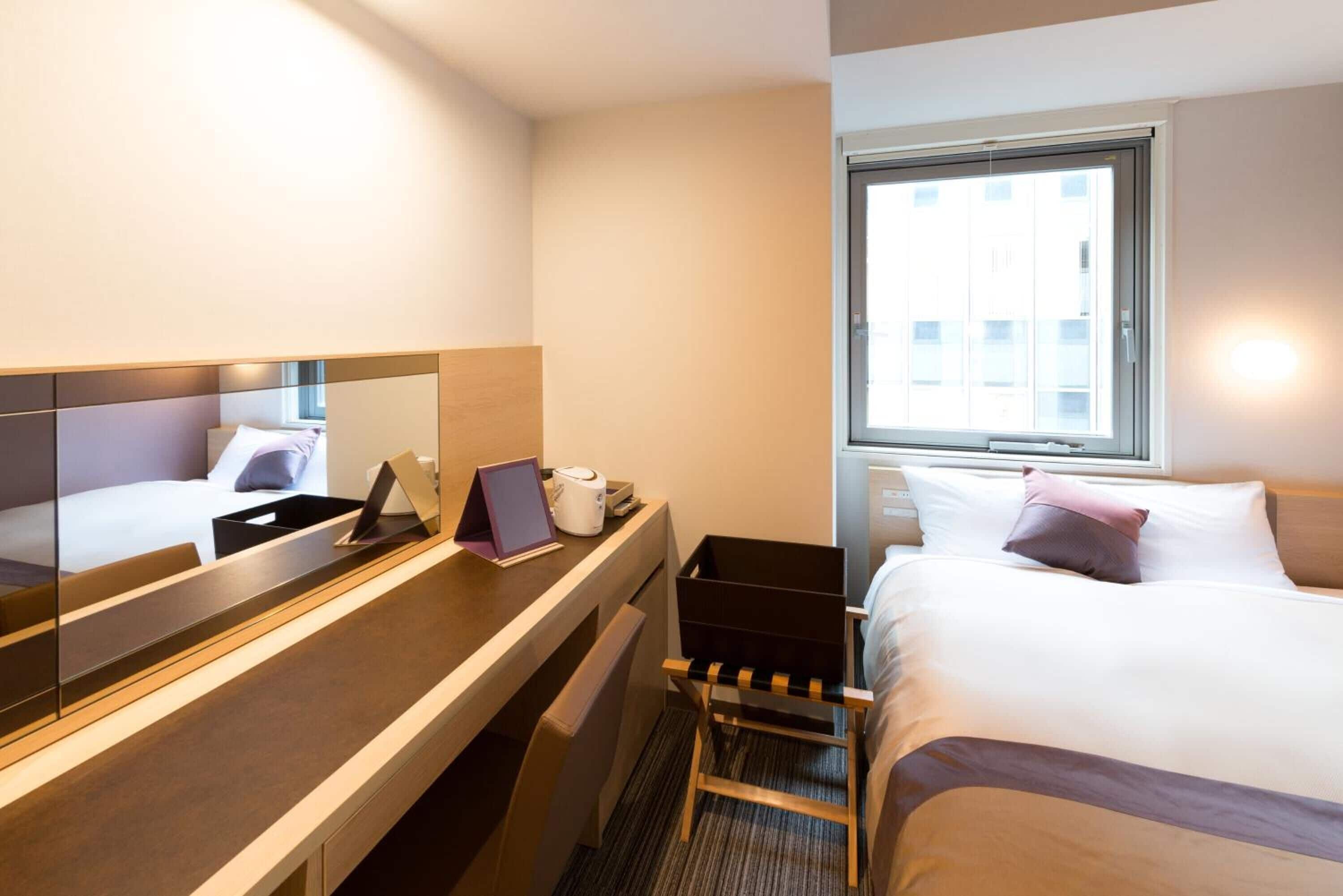 A hotel room with a bed, a desk with a laptop, a chair, a small table, a mirror, and a window.
