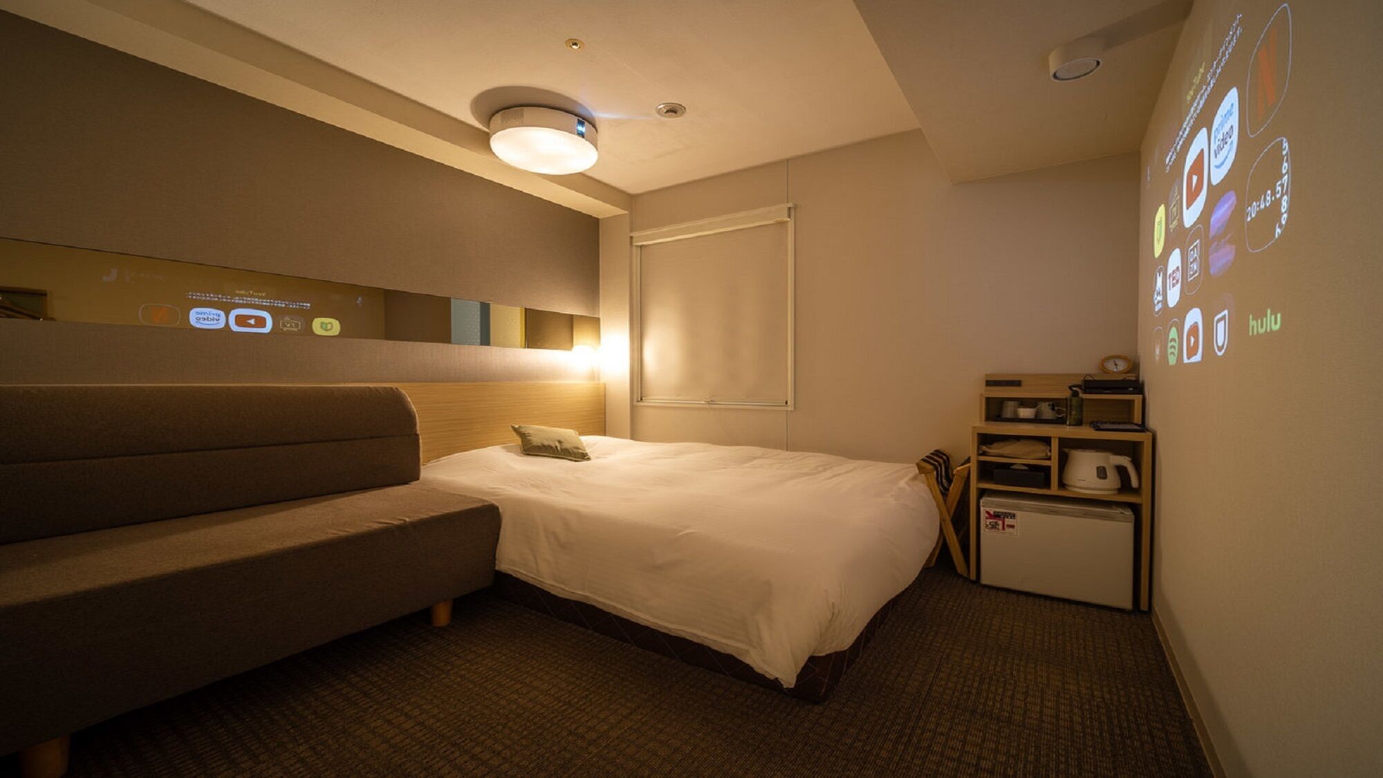 Hotelkamer met een bed, een bank, een klein bureau met een waterkoker en een muur waarop diverse iconen geprojecteerd worden.