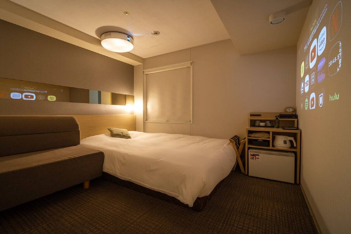 A hotel room with a bed, a sofa, a small desk with a kettle, and a wall with a projection of various icons.