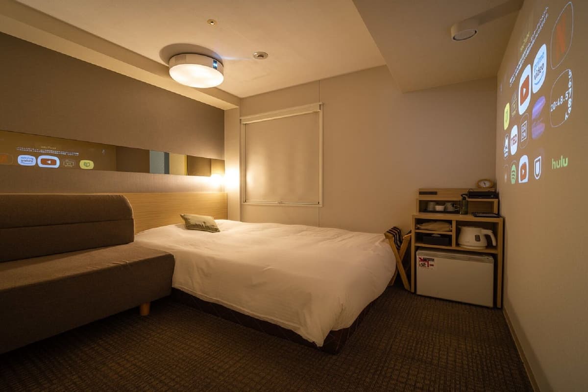 Habitación de hotel con cama, sofá, un pequeño escritorio con hervidor y una pared con proyecciones de varios iconos.