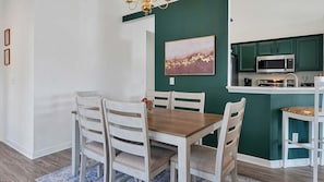 Comedor con mesa de madera y sillas blancas, pared de acento verde, y cocina con armarios color turquesa y un microondas.
