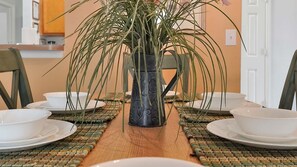 Une table à manger dressée avec de l’assiette et des bols blancs, un vase décoratif contenant des feuilles vertes et des sets de table en fibres tressées.