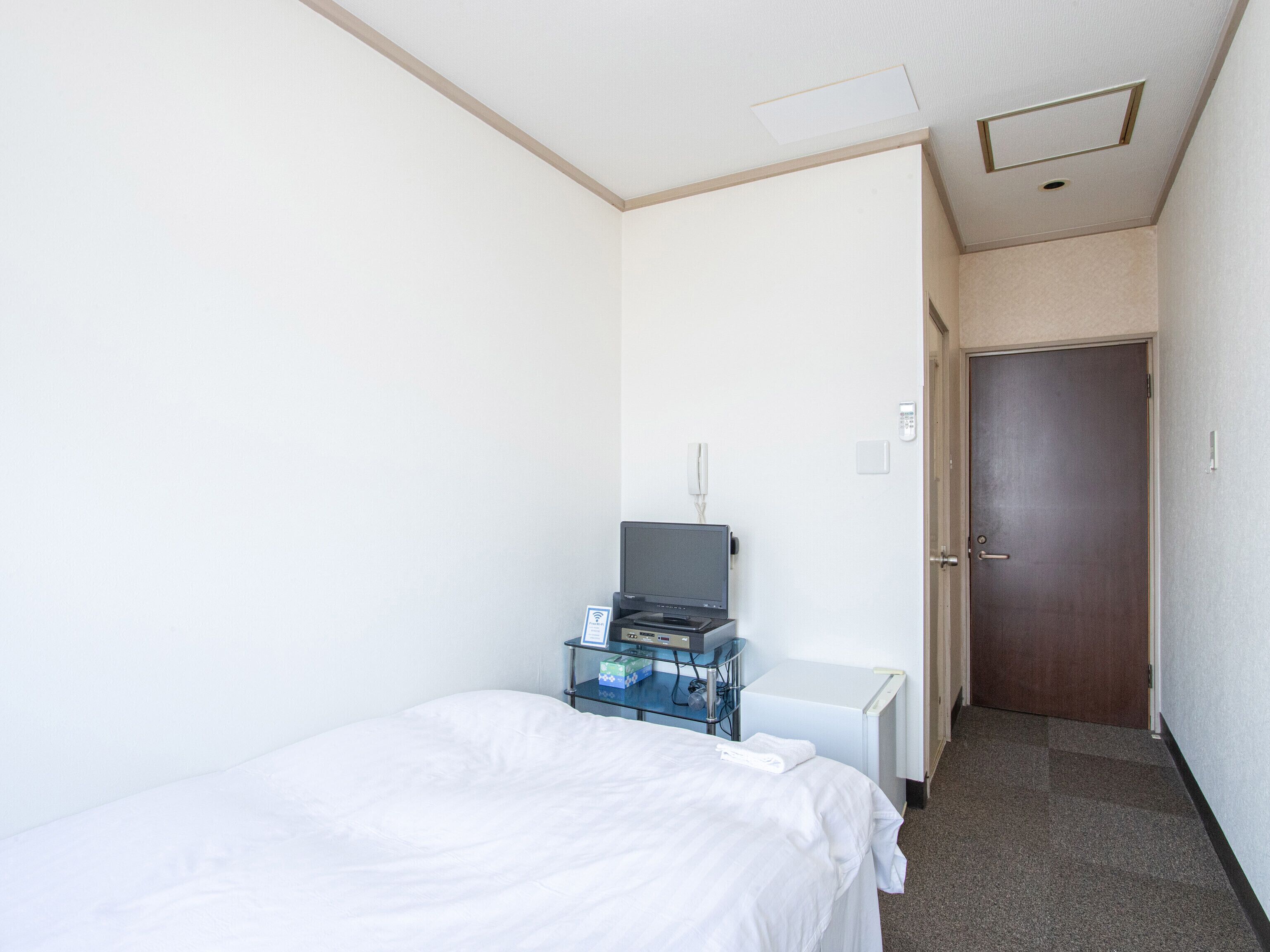 Una camera d'albergo con un letto singolo, un tavolino con la televisione, un comodino con un telefono e una scatola di fazzoletti, e una porta che dà su un'altra stanza.