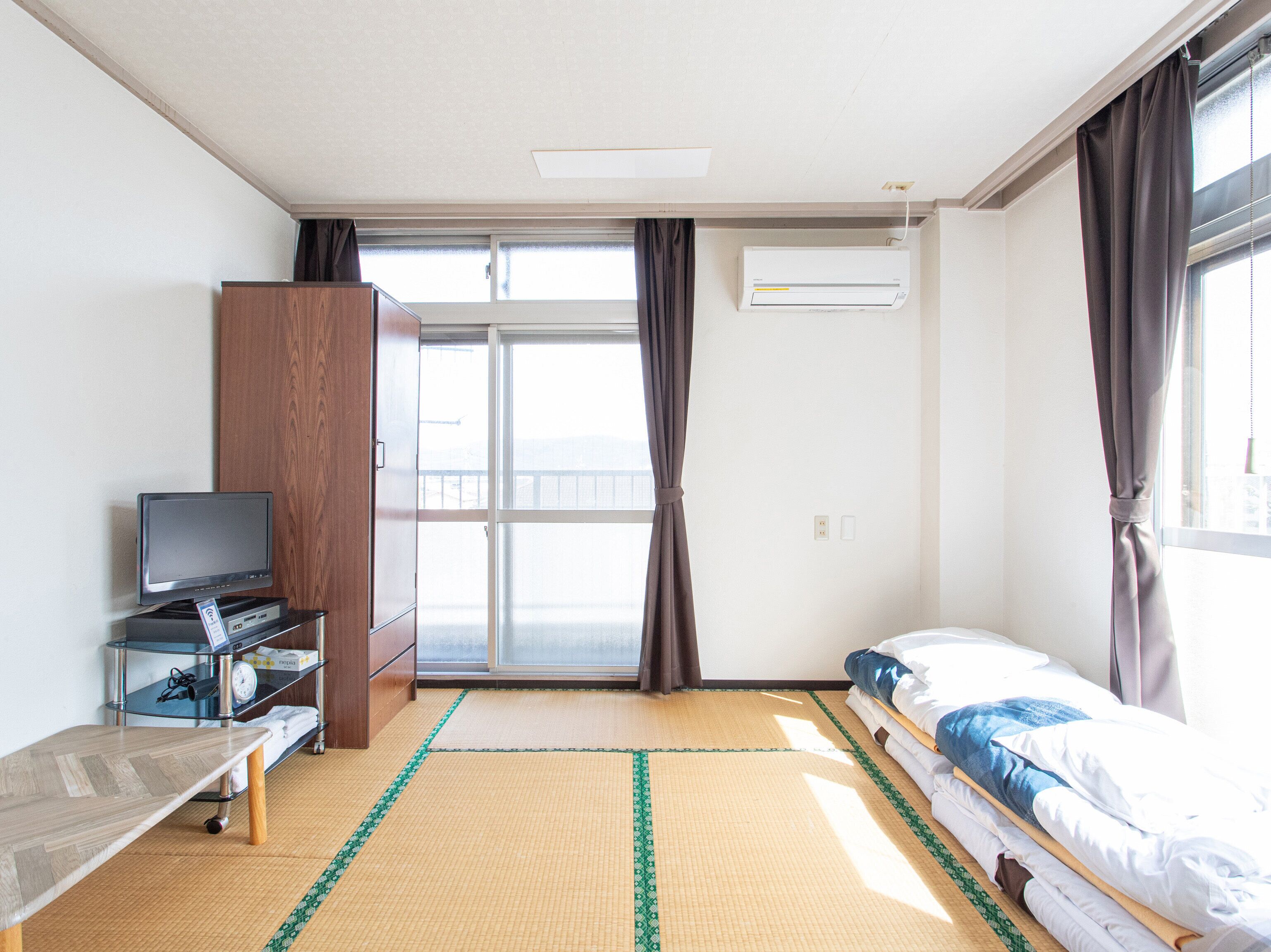 A hotel room with a bed, a TV on a stand, a wardrobe, and a large window with curtains.
