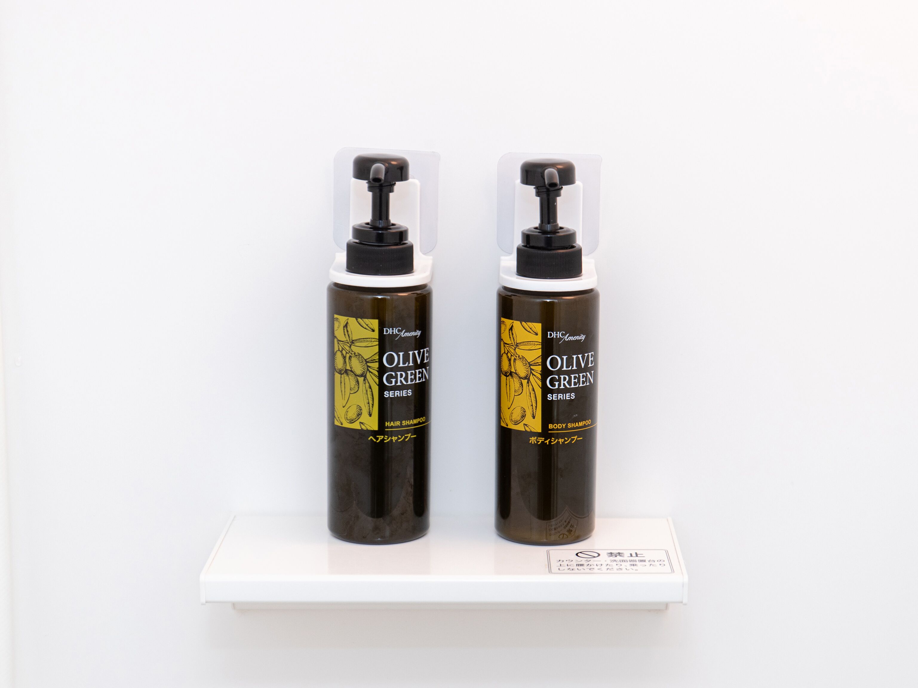 Two bottles of olive green hair and body shampoo on a white shelf.