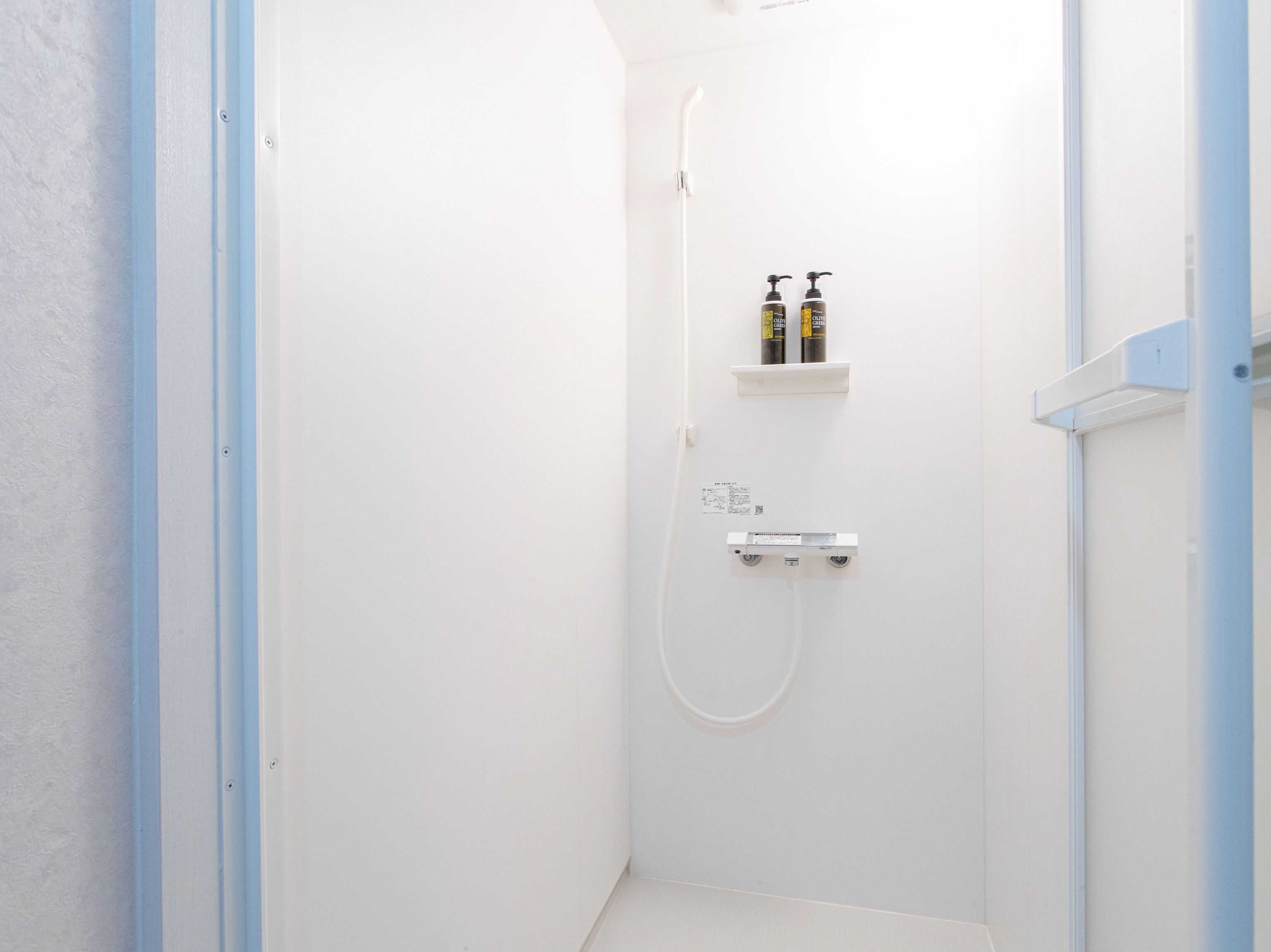 A small, white shower room with a white door, a white showerhead, and two bottles of shampoo on a shelf.
