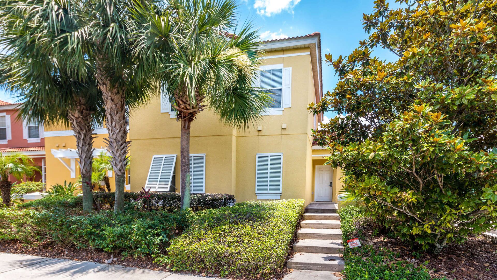 A yellow building with white shutters, surrounded by lush greenery and palm trees.
