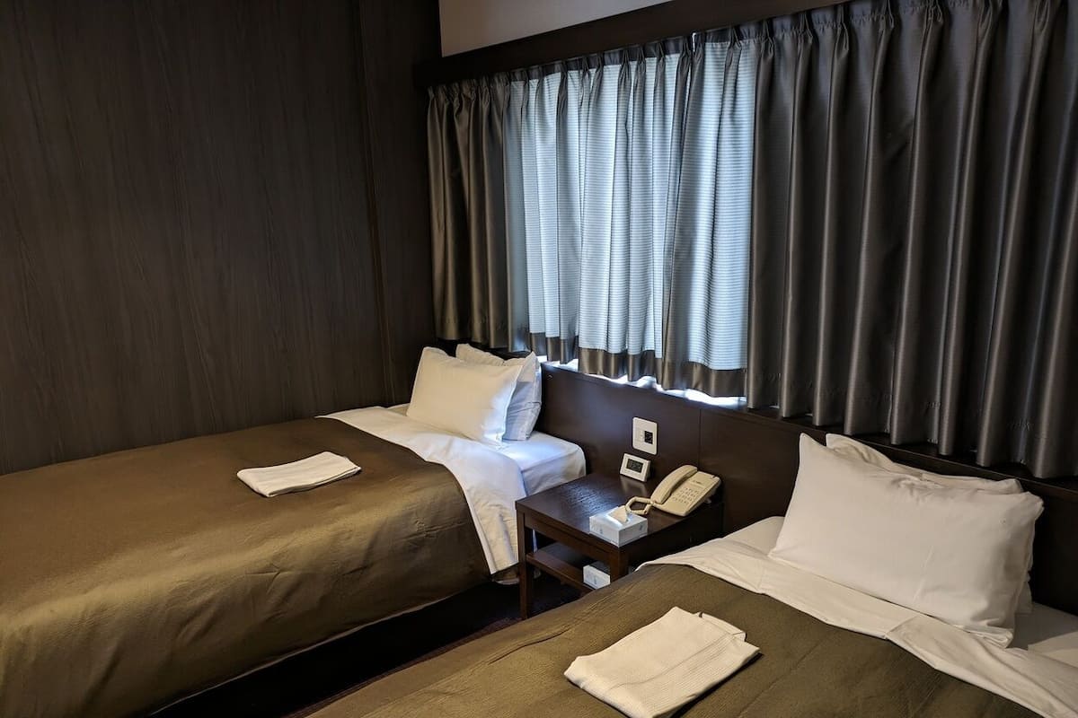 Una habitación de hotel con dos camas, una mesita de noche con teléfono y una ventana con cortinas.