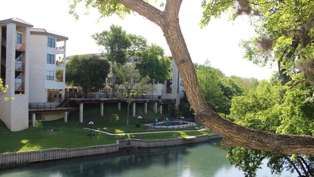 A modern building with a pool, surrounded by lush greenery and a tree. - Blue River Condo on the Comal River (New Braunfels)