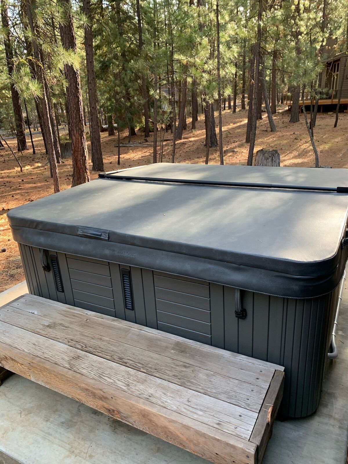 A large, gray outdoor hot tub with a wooden bench in front, surrounded by a forested area.