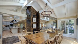 A spacious dining area with a large wooden table and chairs, a chandelier, and a view of the outdoors through large windows. - 12 Royal Tern - 2nd Row (70 yard walk) to the Ocean and Large Private Pool (Hilton Head Island)