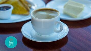 Una taza blanca de café sobre un plato con una bandeja de comida al fondo.