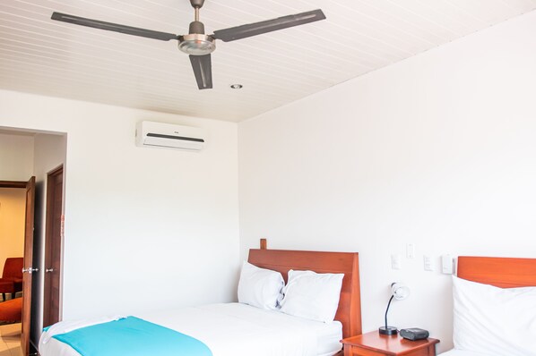 A hotel room with a bed, a ceiling fan, an air conditioning unit, a bedside table with a lamp, and a red chair.
