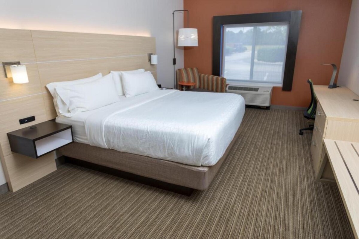 A hotel room with a bed, bedside table, lamp, chair, and a window with a view.