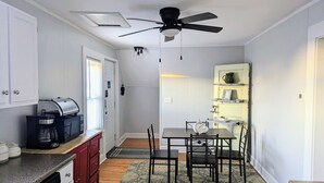 Una cocina compacta con cafetera, una mesa de comedor con sillas, un ventilador de techo y un pequeño estante con objetos decorativos.