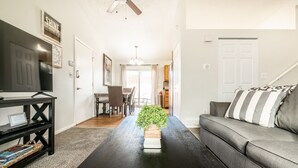 A living room with a black coffee table, a grey sofa, a TV on a black stand, and a dining area with a table and chairs. - "Central Springs" Super Cozy 4Bed / 2Bath Home GREAT Location! Click to see more (Colorado Springs)