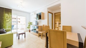 A modern living room with a green sofa, wooden dining table, and chairs. There is a TV on a cabinet, a plant, and a view of the outside.