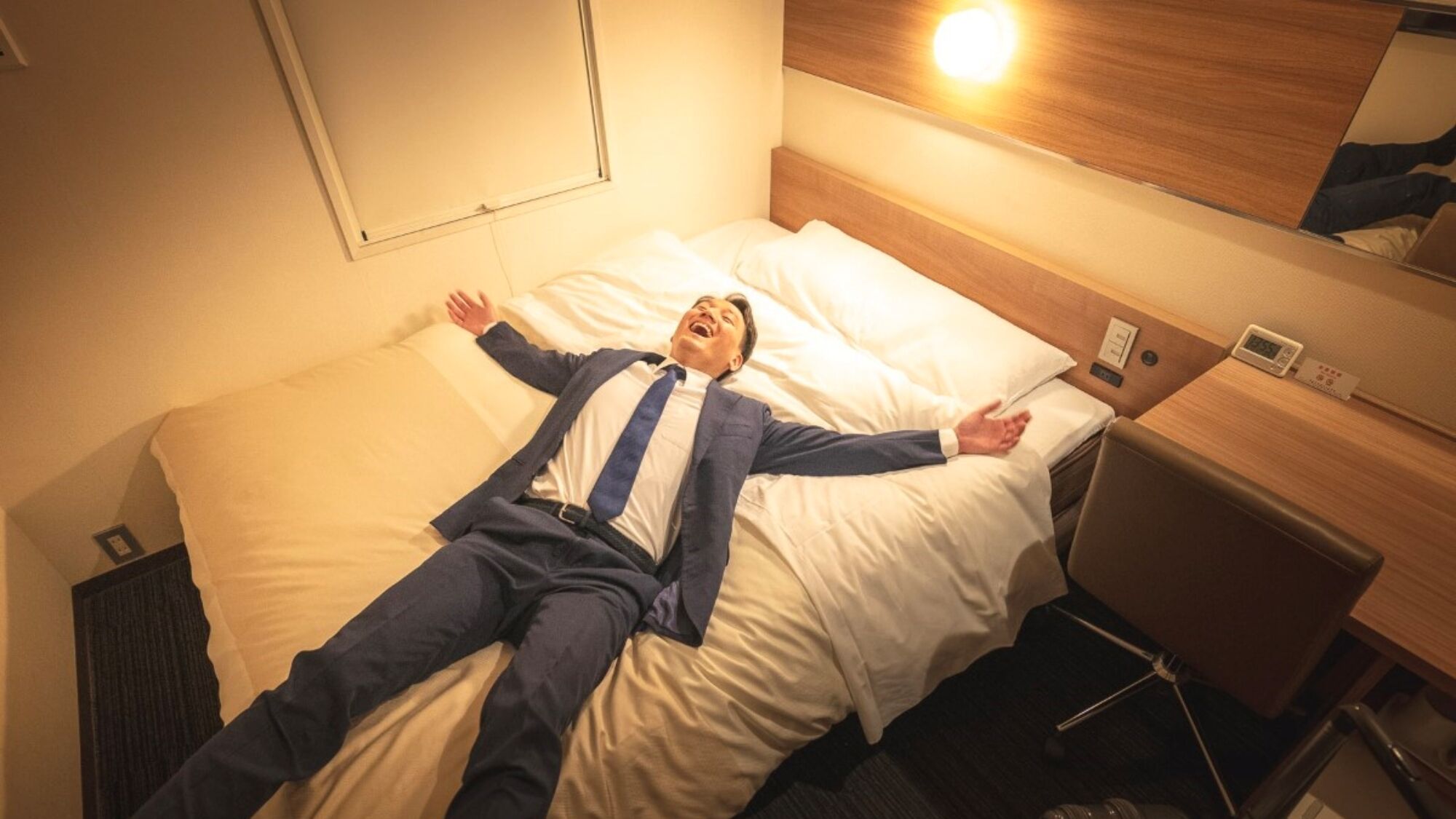 Una habitación de hotel con una cama, una mesita de noche con lámpara, un reloj y un cajón pequeño.