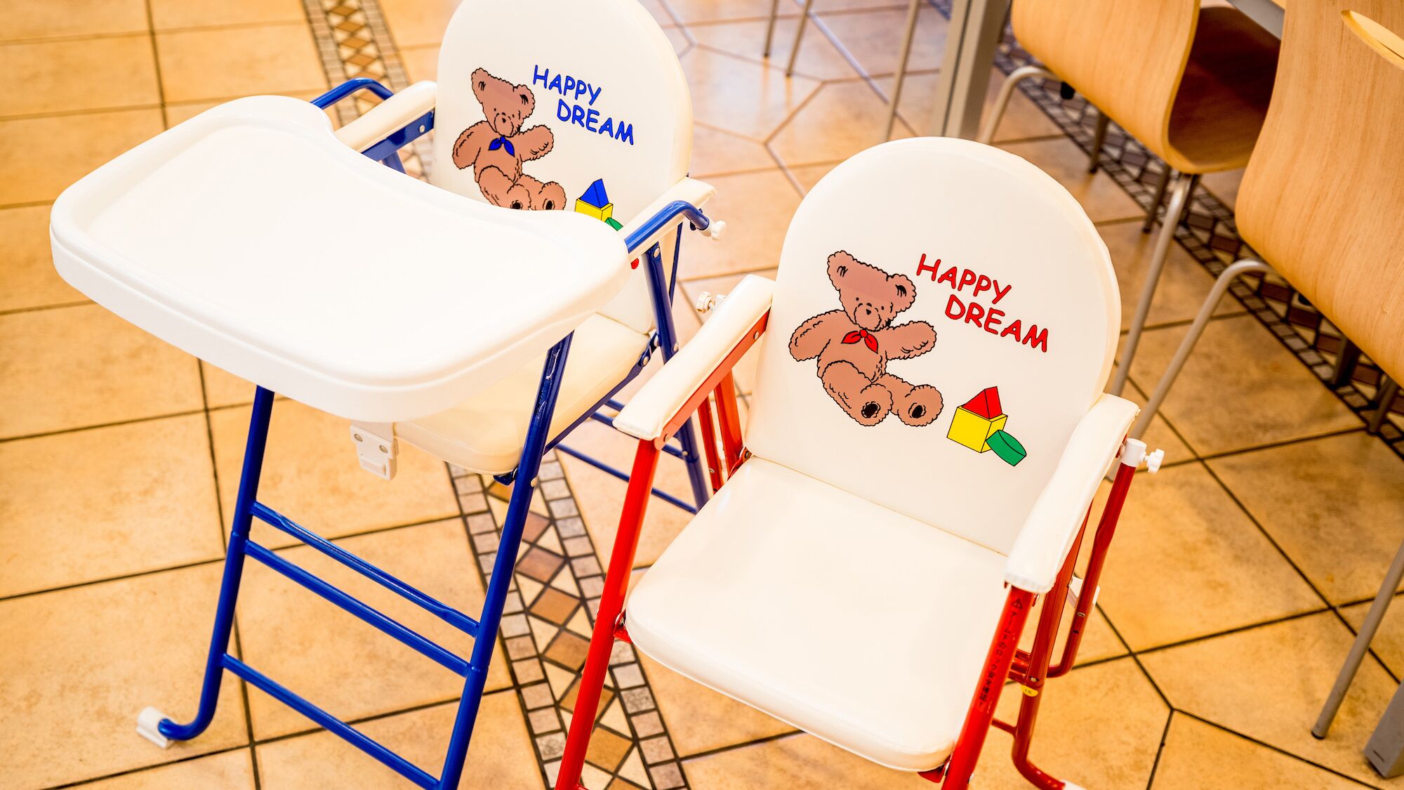 Two high chairs with 'HAPPY DREAM' and a teddy bear design, in a tiled room with a metal chair in the background.