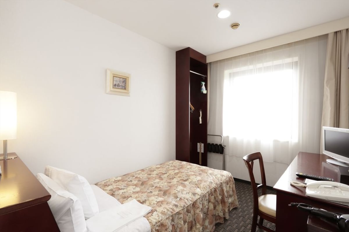 A hotel room with a bed, a desk with a computer and phone, a chair, a lamp, a wardrobe, and a window with curtains.