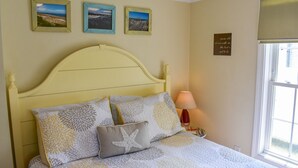 Una cama con cabecera amarilla, varios almohadones y un cojín con forma de estrella de mar. En la pared hay tres cuadros enmarcados y una lámpara de noche.