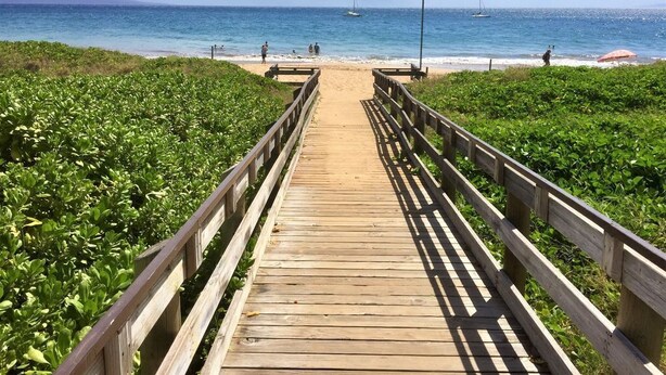 Una pasarela de madera que lleva a una playa con barcos a lo lejos.