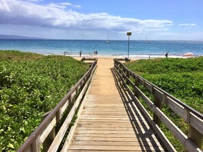 Ein hölzerner Steg führt zu einem Strand, im Hintergrund sind Boote zu sehen.