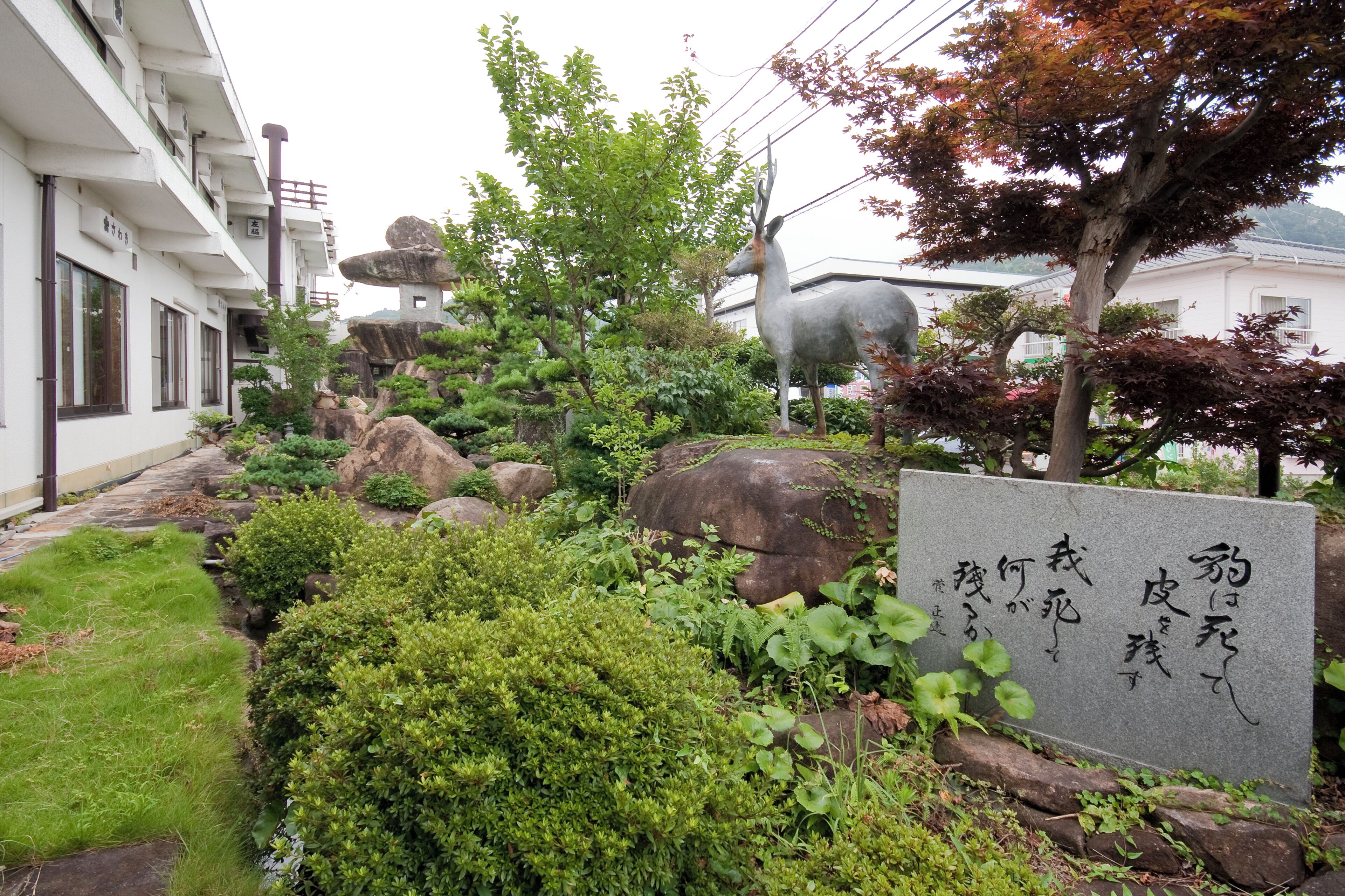 Un jardín ajardinado con una estatua de ciervo, rocas y una lápida de piedra con caracteres japoneses.