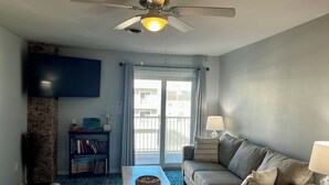 A living room with a ceiling fan, a sofa, a coffee table, a bookshelf, and a view of the outside through a sliding door.