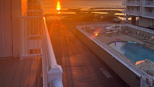 A balcony with a view of the sunset over the ocean, a pool area with lounge chairs, and a multi-story building in the background.