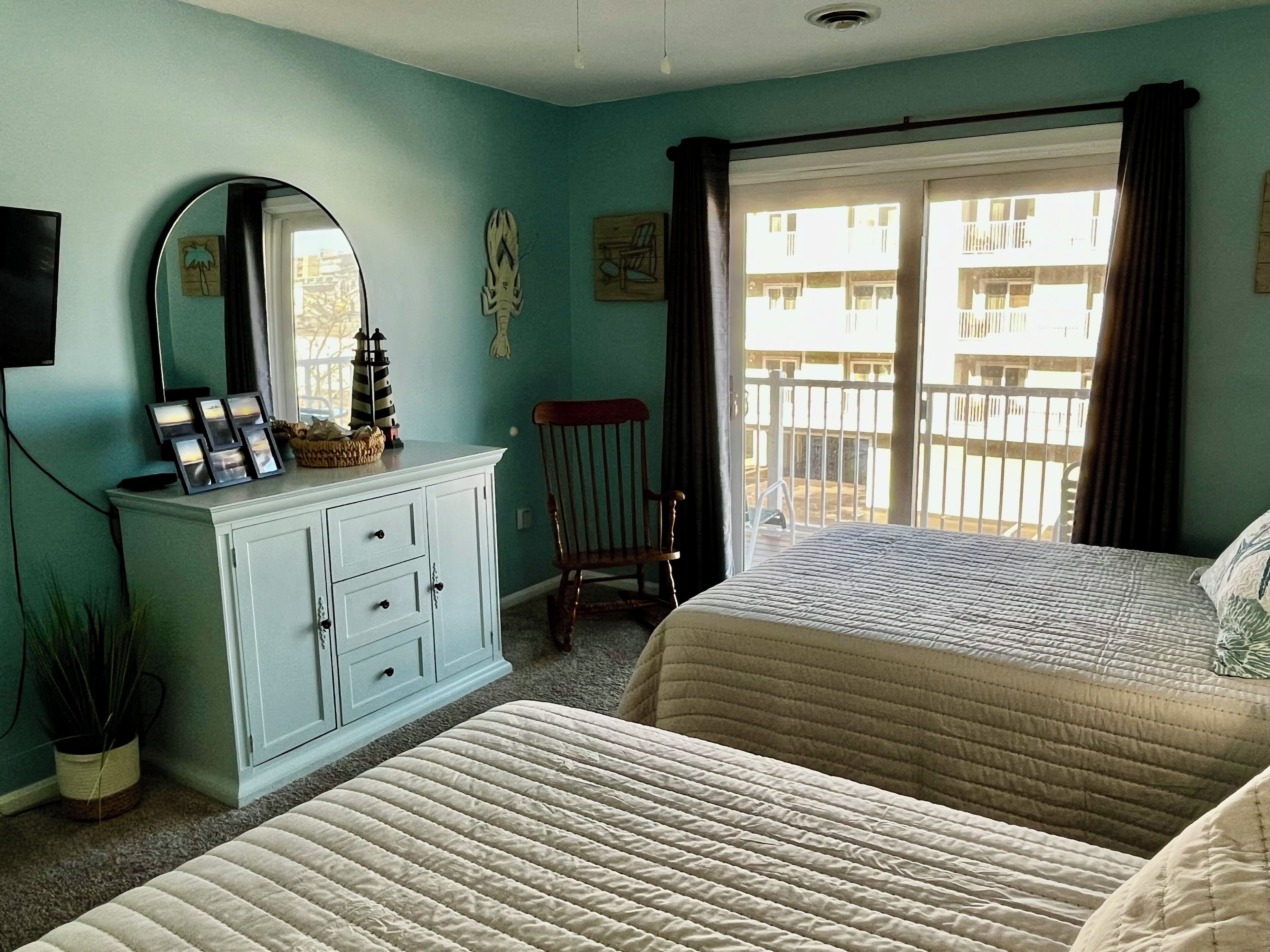 A bedroom with two beds, a dresser, a mirror, and a window with a view of buildings.