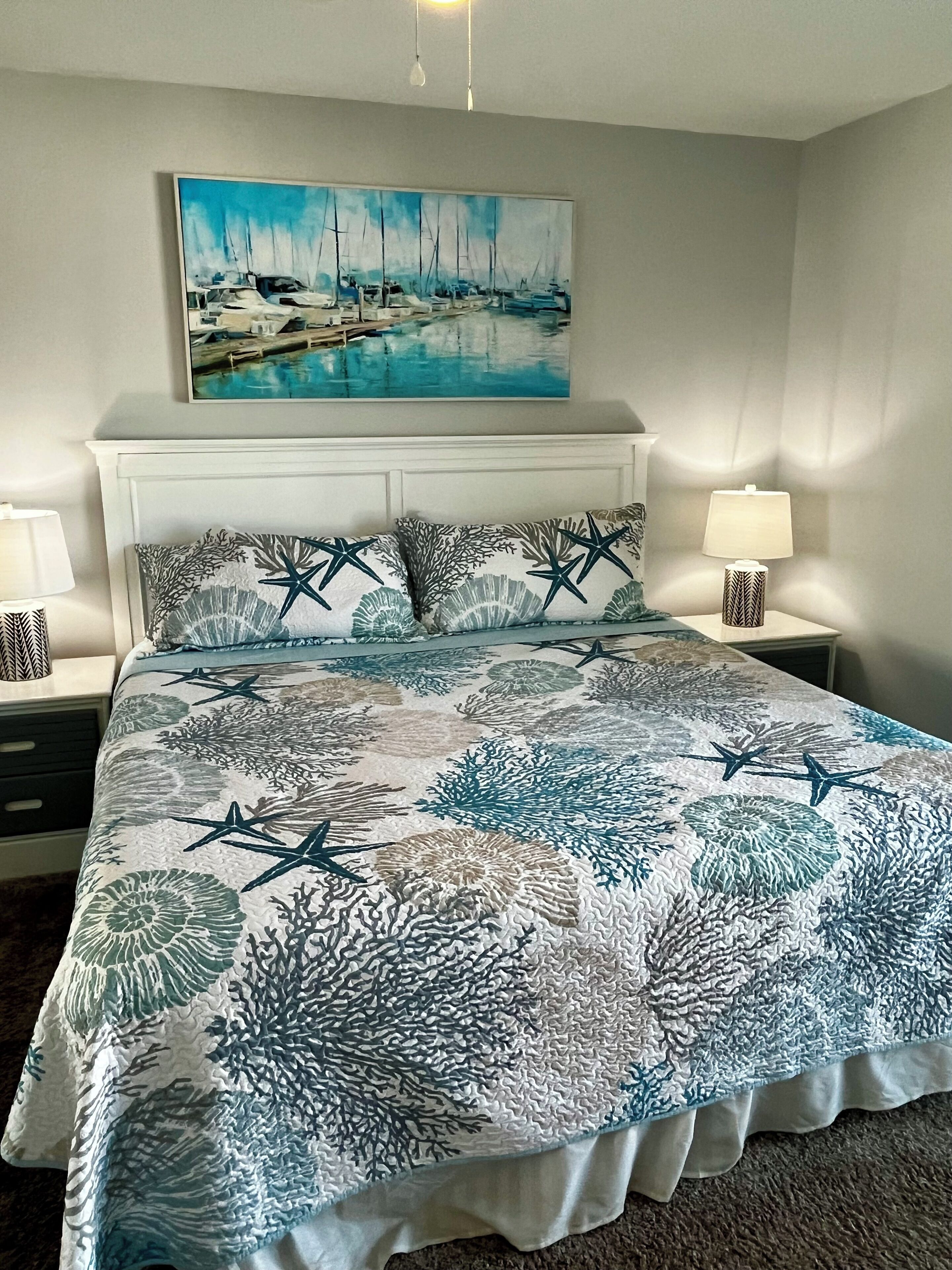 A bedroom with a bed, two bedside tables, a nightstand, a painting above the bed, and a window with curtains.