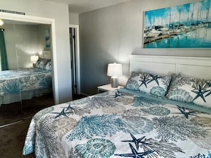 Une chambre d’hôtel avec un lit orné d’un motif représentant des étoiles de mer et des coraux, une table de chevet avec une lampe, un tableau représentant un port de plaisance et une porte donnant sur une autre pièce.