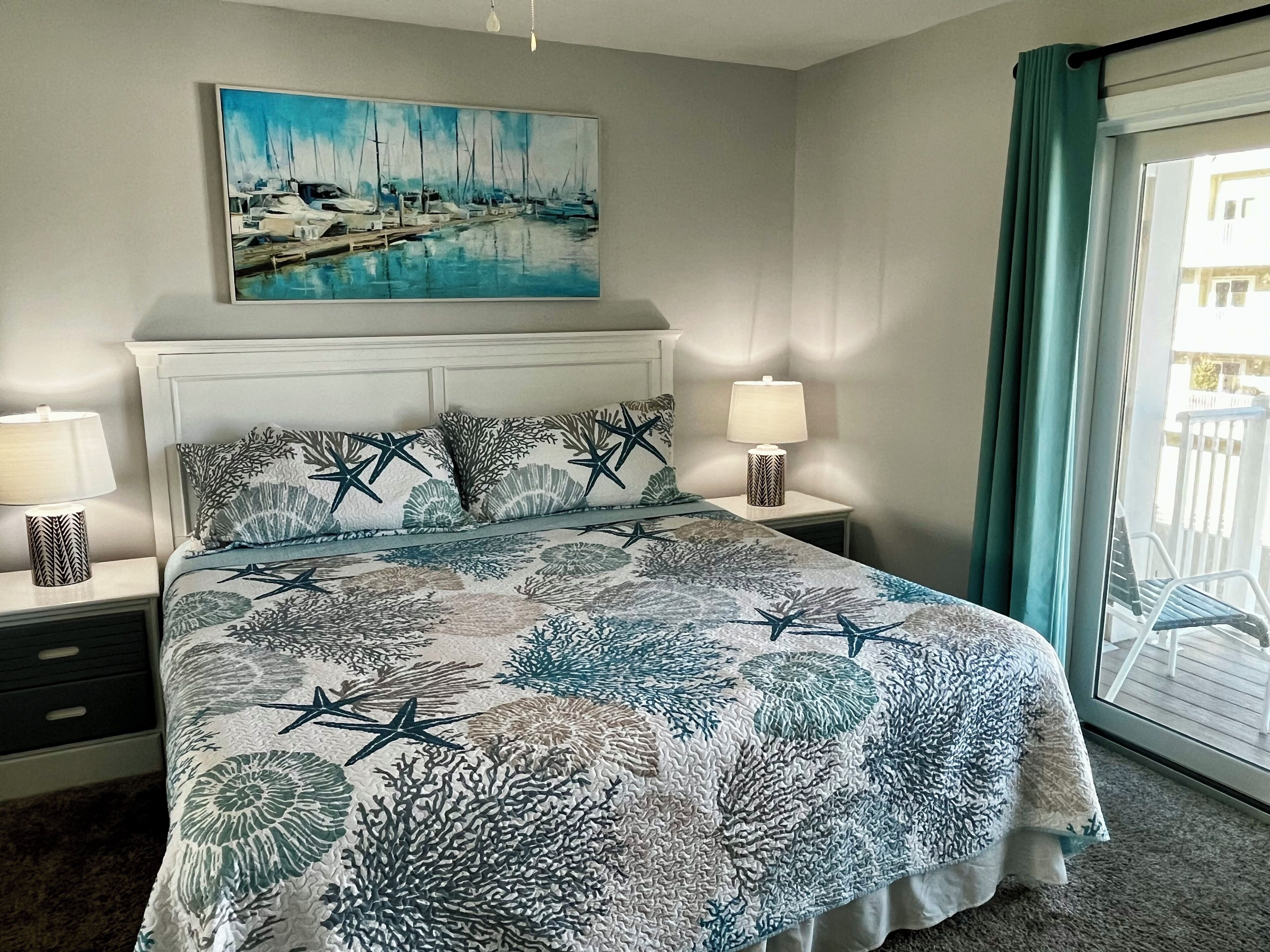 A bedroom with a bed, two bedside tables with lamps, a painting above the bed, and a view through a window.